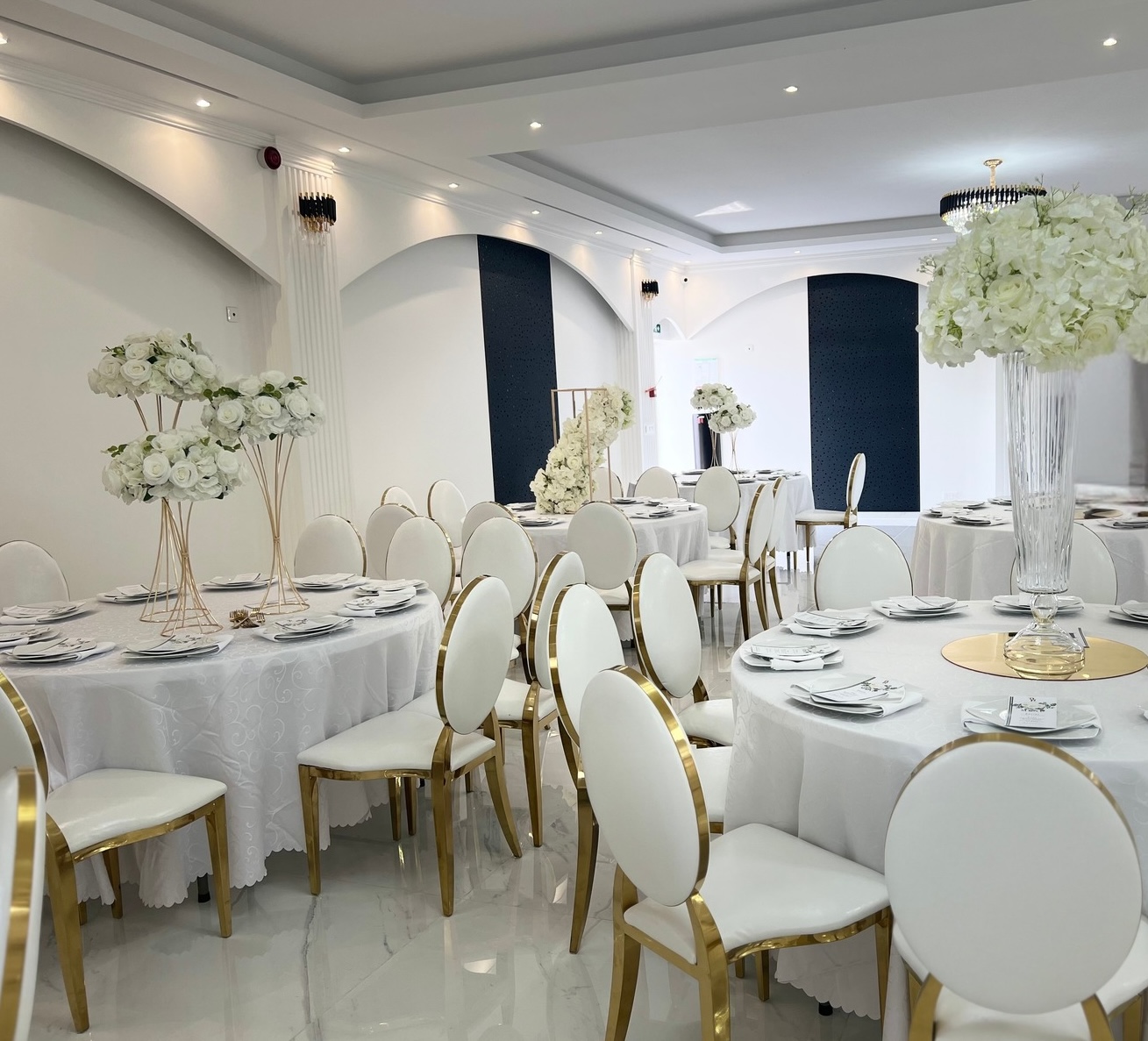 Salle de réception Palace Victory Events à Tremblay-en-France - Tables rondes avec chaises dorées et blanches, centres de table floraux avec roses et hortensias blancs, lustres en cristal